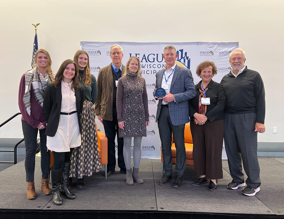 A group of community leaders stand in front of a sponsor board holding an award